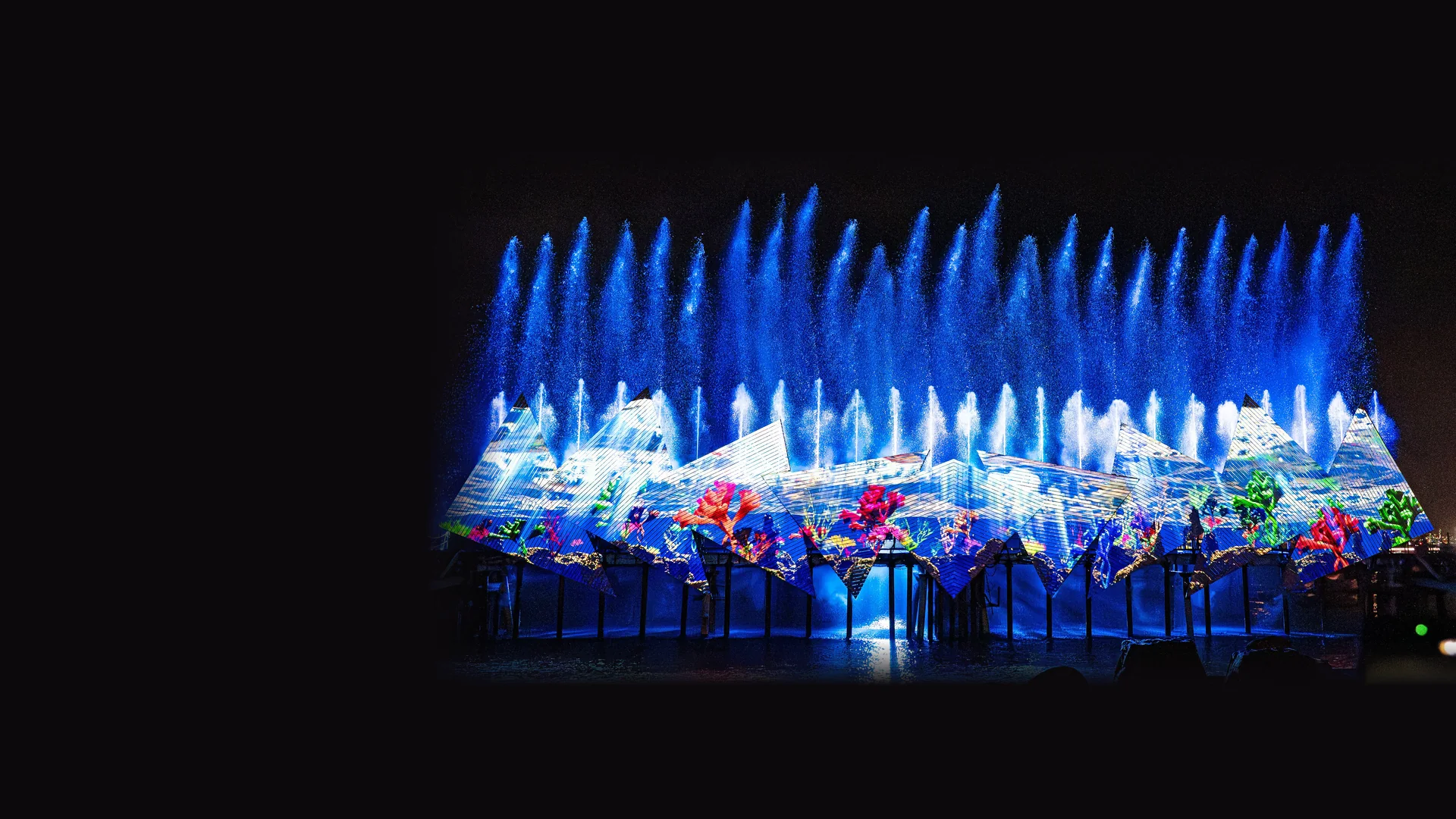 Water jets rise in front of a large angular projection screen showing a coral reef scene on the sea at Siloso Beach, Sentosa, at night