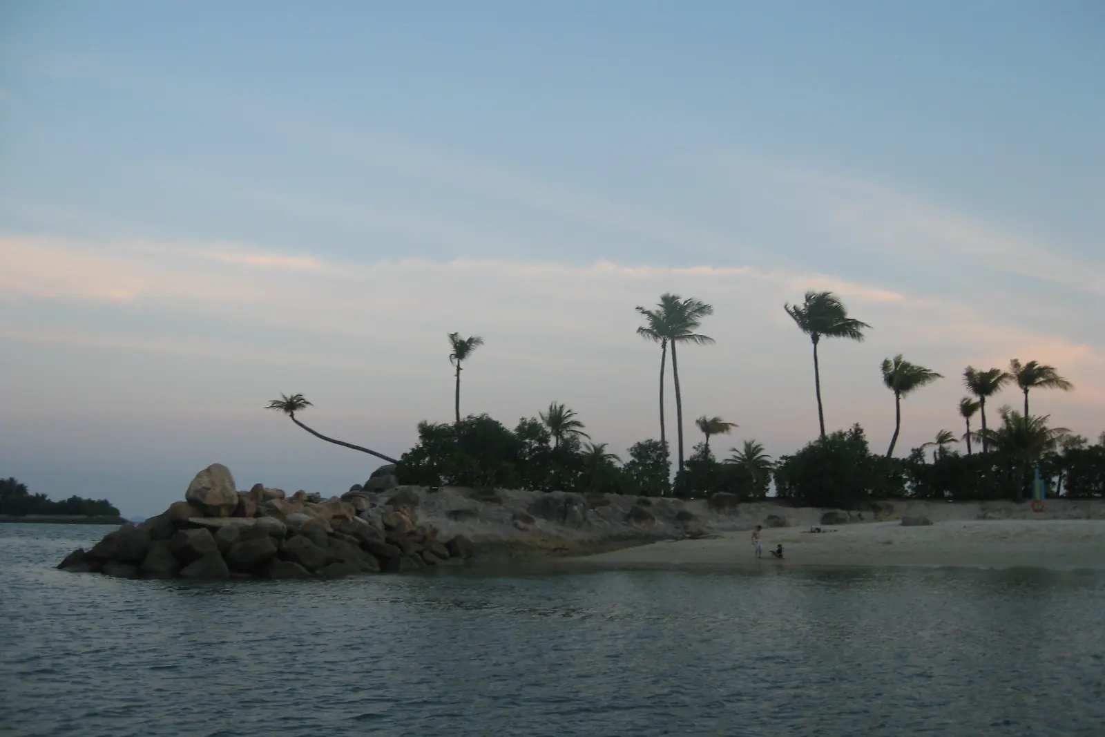 Siloso Beach at dusk, with palm trees along a rocky point and a pale pink and blue sky over the water