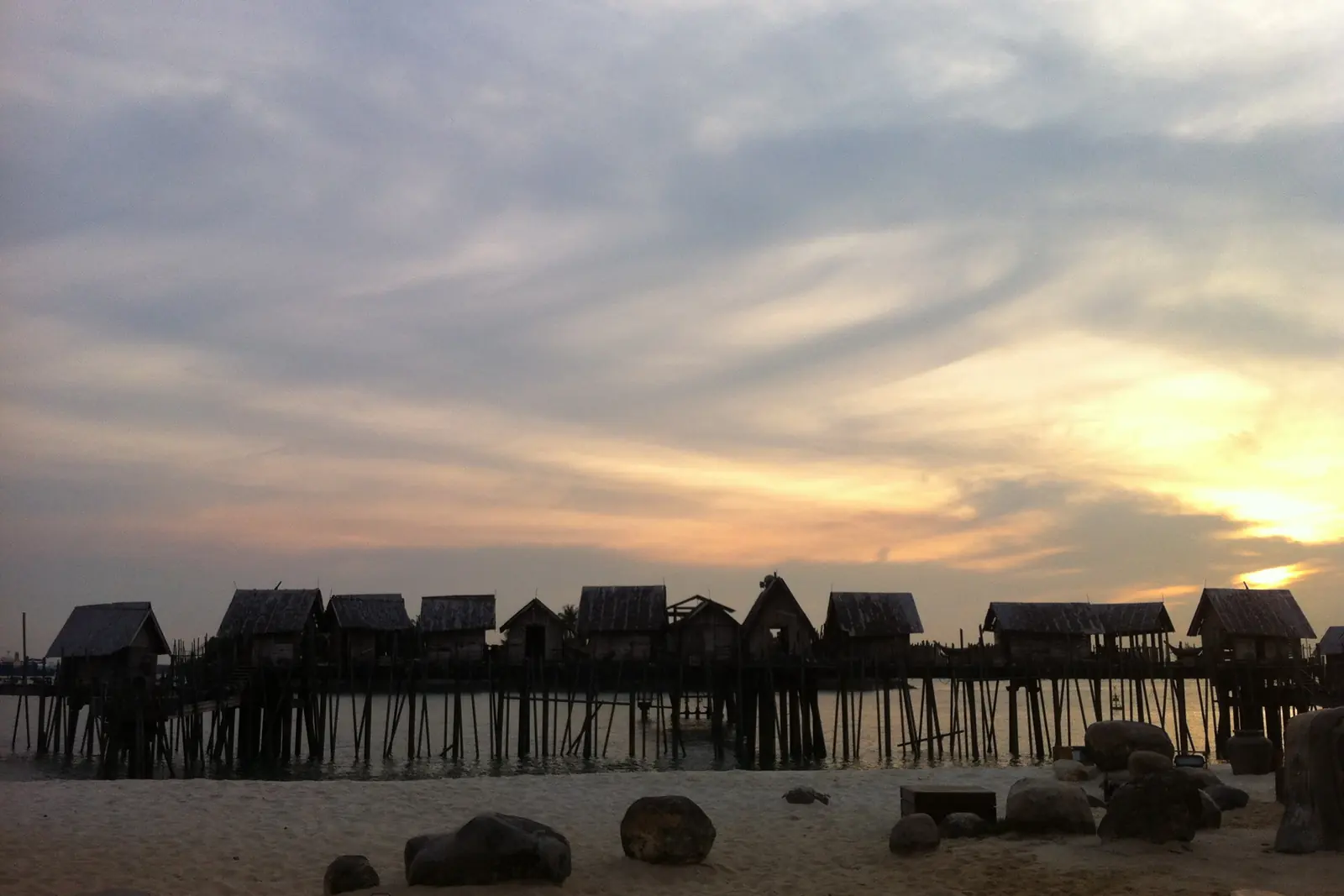 The stage of Songs of the Sea, the show that Wings of Time replaced, with a row of stilt huts silhouetted against a sunset sky on Siloso Beach