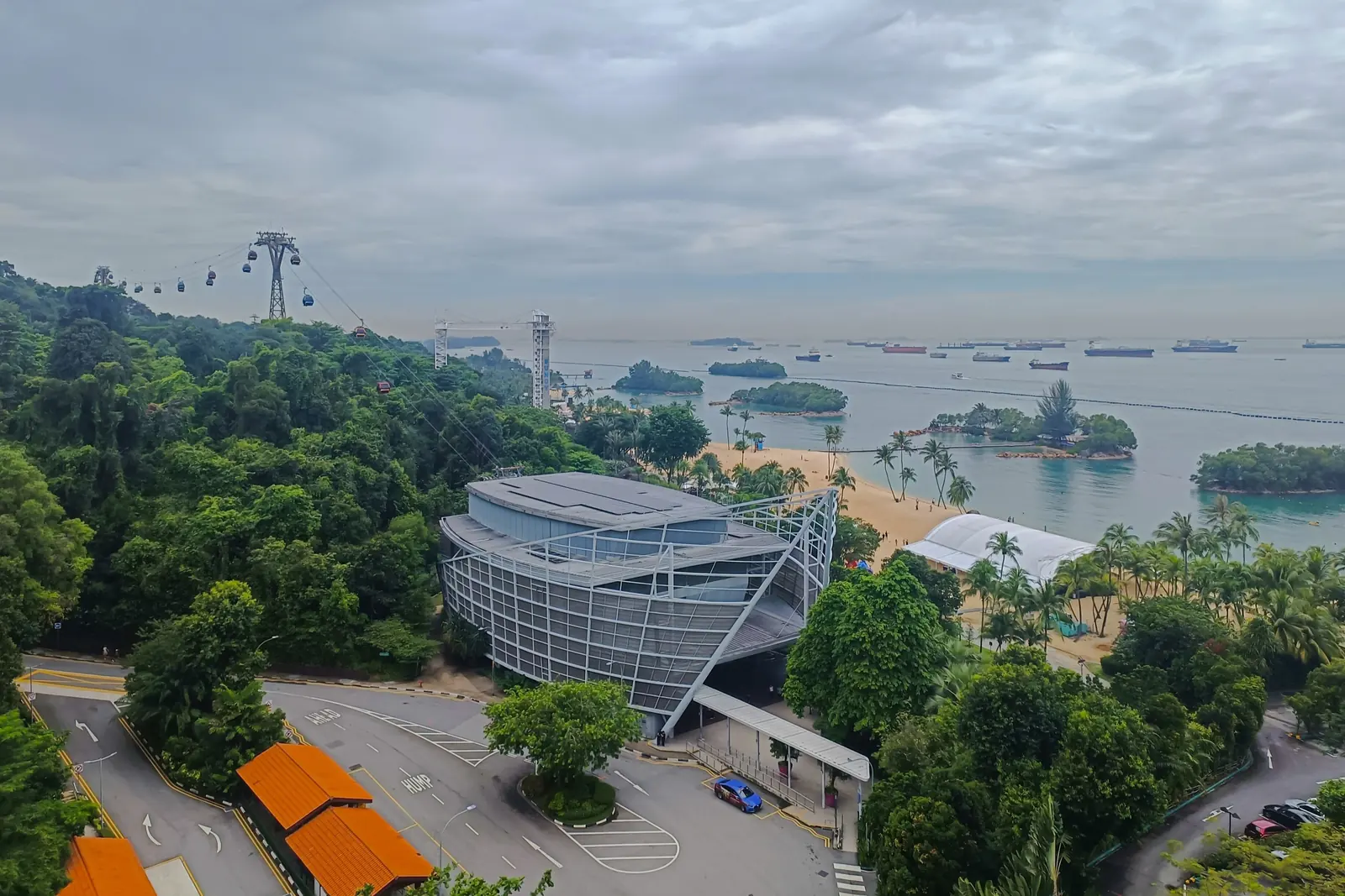 A view over part of Sentosa with cable car cabins on their line above the trees and the sea and ships on the horizon